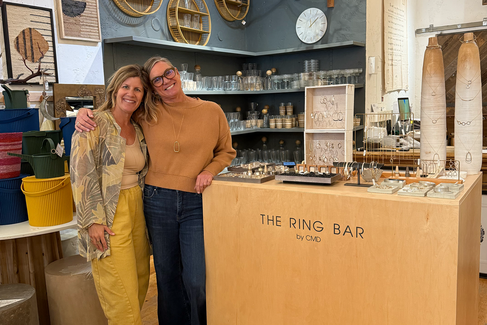 Colleen Mauer smiling with a friend behind The Ring Bar by CMD jewelry display at Abode, surrounded by handcrafted jewelry, home decor, and curated accessories.